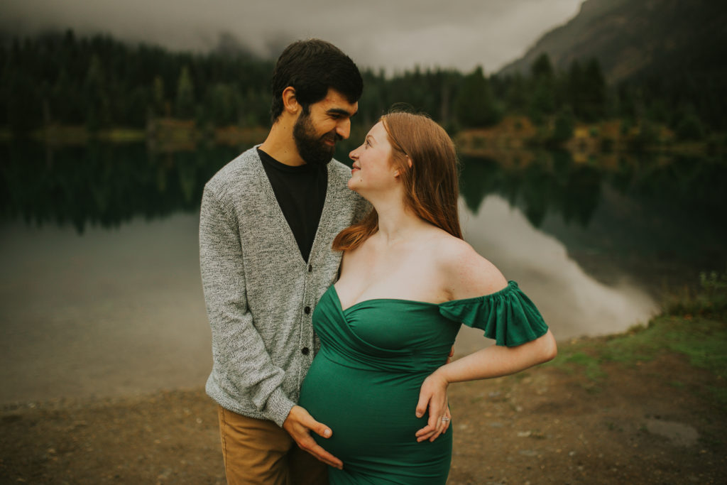 Man in grey cardigan holding pregnant wife's belly who is wearing a green off shoulder dress