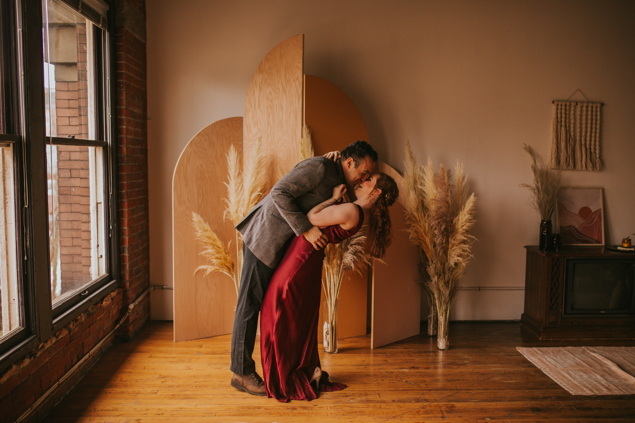 bride and groom dip into a kiss in front of the wedding arch in a photo studio bride is wearing a read dress and groom is wearing a grey tuxedo