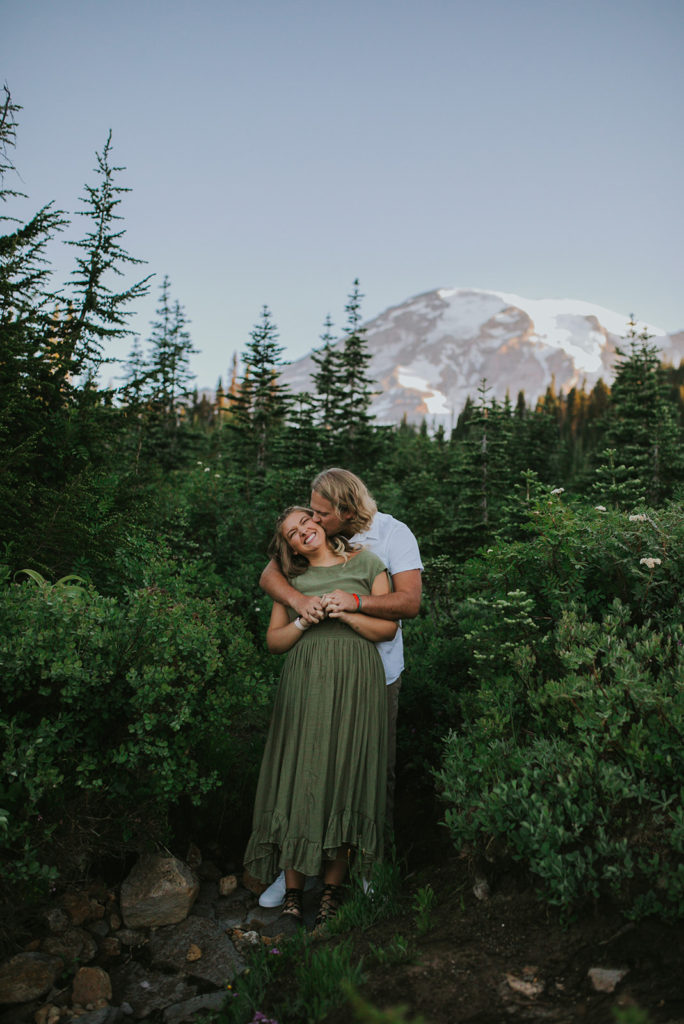 Couple standing in a forest with a mountain in the background. Male partner is standing behind the female partner and hugging from behind. Male partner is kissing female partner on the cheek while the female partner smiles.