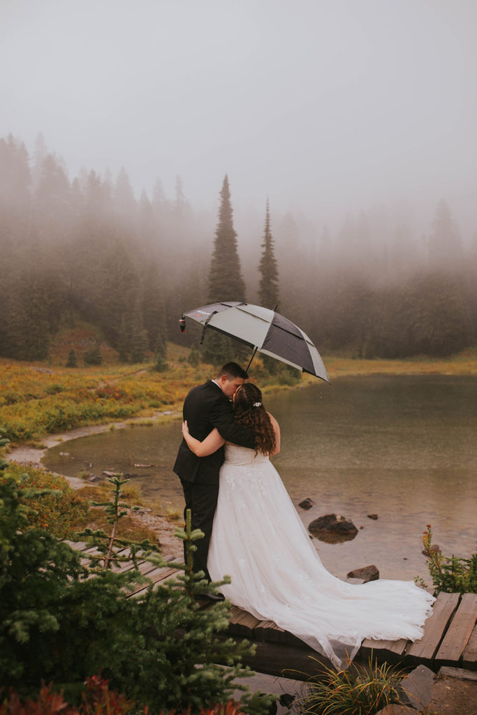 Wedding couple standing on a bridge holding an umbrella while it rains and kissing while hugging each other